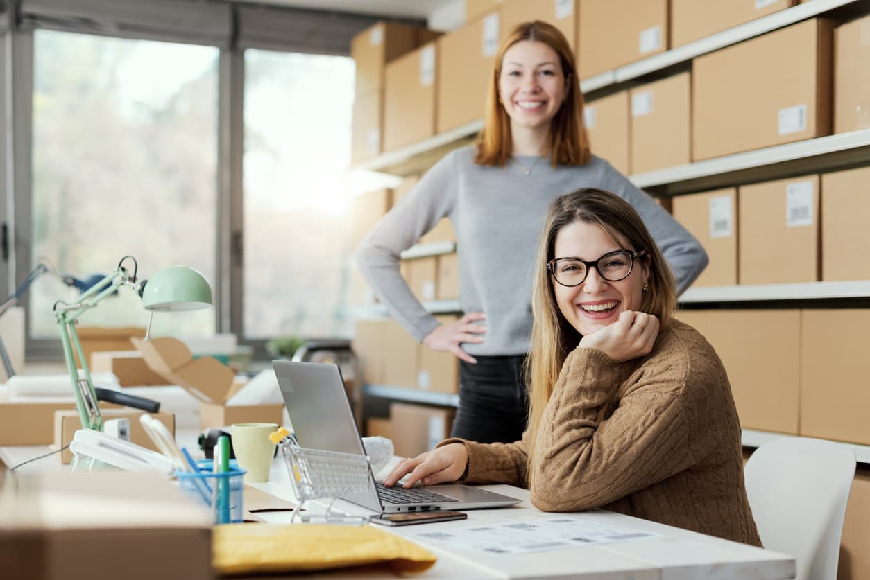 Two small business owners smiling in their fulfillment room, one working at a laptop surrounded by shelves of packed orders